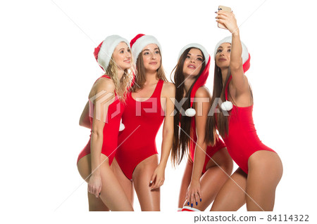 Happy young millennial women have fun celebrating Christmas holiday taking photos and selfies using a mobile phone posing in red swimsuits and Santa hats isolated on white background 84114322