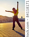 sporty athletic woman with dyed pink hair stands back, stretches against background of sunset 84118171