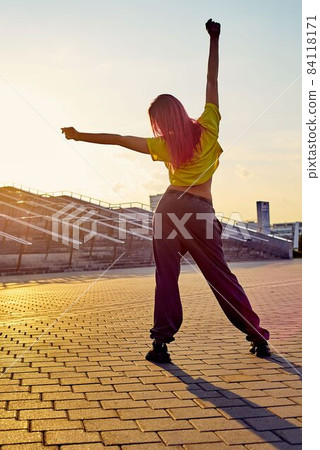 sporty athletic woman with dyed pink hair stands back, stretches against background of sunset 84118171