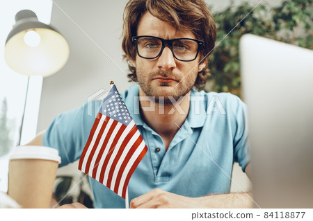 Young bearded businessman sitting at table with laptop and american flag Young bearded businessman sitting at table with laptop and american flag 84118877