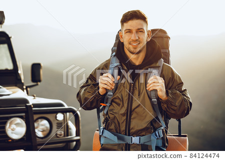 Portrait of a young traveler man in hiking equipment standing near his off-road car Portrait of a young traveler man in hiking equipment standing near his off-road car 84124474