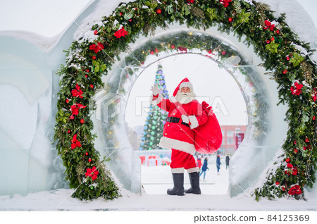 Santa Claus posing with a bag of gifts on the background of Christmas decorations outdoors Santa Claus posing with a bag of gifts on the background of Christmas decorations outdoors 84125369