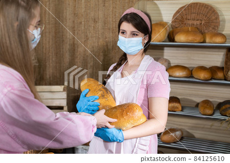 A customer buys more loaves of bread for her family. The saleswoman serves the bread. A saleswoman during the COVID-19 pandemic is wearing gloves and a face mask at a bakery store. A customer buys more loaves of bread for her family. The saleswoman serves the bread. A saleswoman during the COVID-19 pandemic is wearing gloves and a face mask at a bakery store. 84125610