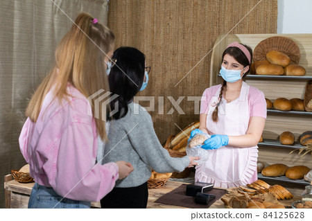 A saleswoman in a protective mask serves bought bread by two teenagers. Sales during the ongoing COVID-19 pandemic. A saleswoman in a protective mask serves bought bread by two teenagers. Sales during the ongoing COVID-19 pandemic. 84125628