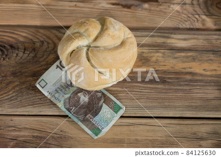A round bun with partitions and cracks is lying on an old wooden table and a banknote is under the bun. Bun on the background of old boards. Ten Polish zlotys. A round bun with partitions and cracks is lying on an old wooden table and a banknote is under the bun. Bun on the background of old boards. Ten Polish zlotys. 84125630