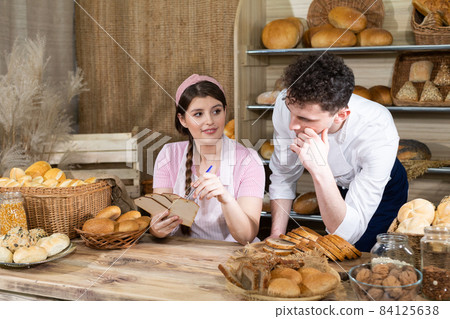 An attractive new saleswoman describes wooden plates with prices for individual types of bread under the supervision of a bakery owner, a young handsome baker. 84125638