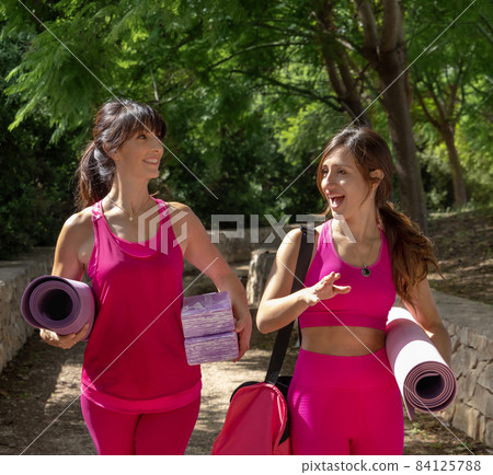 Two women walking in a park with rolled pilated mats and talking Two women walking in a park with rolled pilated mats and talking 84125788