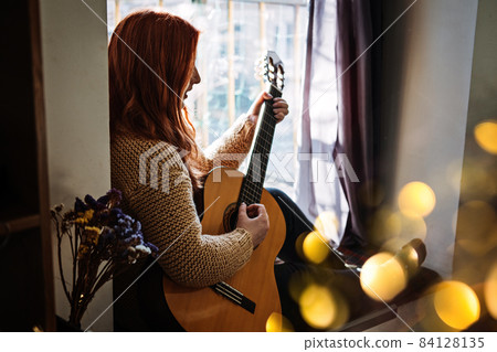 Unaltered candid portrait of young red haired woman in sweater playing acoustic guitar sitting by window at home. Hobbies, indoor home activities for adults in winter, autumn. 84128135