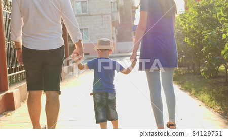 a little boy holding his mother and father's hands in the rays of the sun, walking around the city with the child, a happy family, mom, dad and kid in glare of the sun, childhood dream of baby's life 84129751