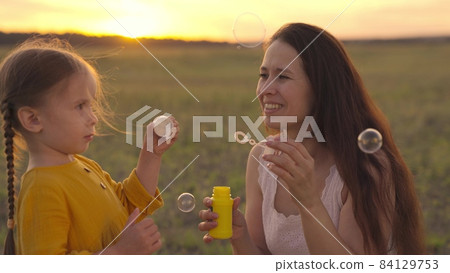 Happy Mother and her little Daughter playing together outdoor, blowing soap bubbles, having fun on backyard. Nature. Beauty Mum and her Child in Park together. Family, Mother's Day Joy. Mom and Baby Happy Mother and her little Daughter playing together outdoor, blowing soap bubbles, having fun on backyard. Nature. Beauty Mum and her Child in Park together. Family, Mother's Day Joy. Mom and Baby 84129753