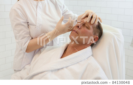 Beautician injecting a senior man into the glabellar region to prevent early signs of aging and expression lines. Mature man receiving beauty injection and anti-aging treatments at a modern spa clinic 84130048