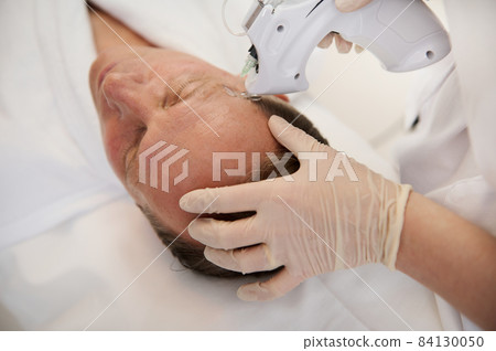 Overhead view of a handsome aged man lying on massage table during mesotherapy gun therapy in modern spa clinic. Anti-aging, rejuvenation and alternative beauty treatment to prevent first wrinkles 84130050