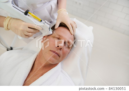 Therapist doing facial mesotherapy treatment on middle aged handsome Caucasian man in a modern wellness spa center. Man getting injections for face tightening at cosmetology clinic 84130051