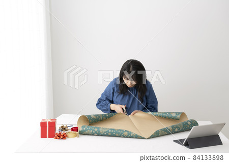 Young woman cutting wrapping paper with scissors to pack gifts at the desk 84133898