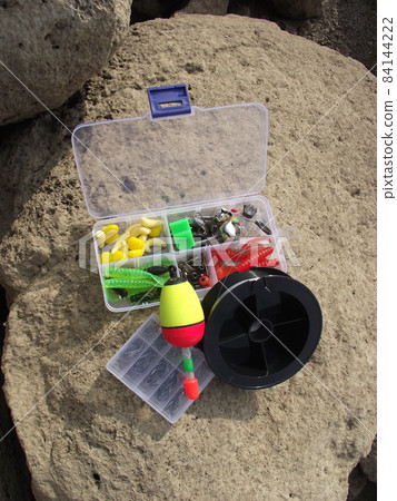 open boxes of translucent plastic with fishing tackle floats and a reel of line lie on a large stone. 84144222