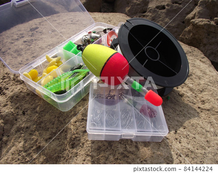 open boxes of translucent plastic with fishing tackle floats and a reel of line lie on a large stone. open boxes of translucent plastic with fishing tackle floats and a reel of line lie on a large stone. 84144224