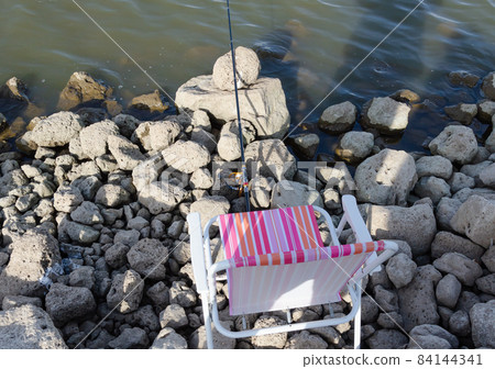 on the rocky shore of the reservoir there is a fishing rod with a reel and a folding chair next to it 84144341
