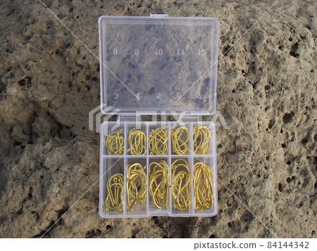 Set of gold-colored fishhooks lies in an open box on stone. 84144342