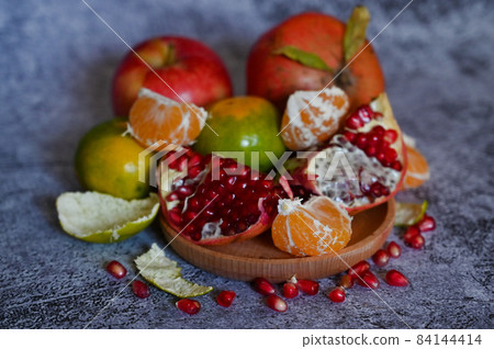 A pomegranate, whole and broken into pieces with other fruits, lies on a marble table 84144414