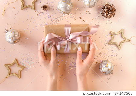 Female hands with natural manicure holding present in kraft paper with pink ribbon on soft beige background. Xmas composition. Flat lay. Happy holidays, New Year celebration and giving love concept.  84147503