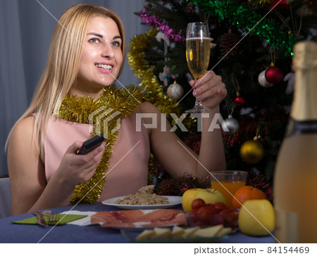 Young female is watching TV and sitting with champagne in New Year night Young female is watching TV and sitting with champagne in New Year night 84154469
