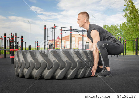 Muscular bearded tattooed fitness man moving large tire in street gym. Muscular bearded tattooed fitness man moving large tire in street gym. 84156138