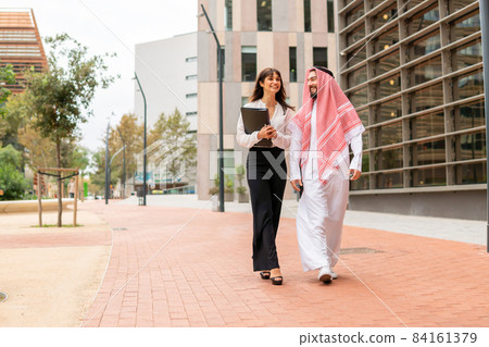 Smiling arab businessman in traditional clothes walking city street with his young attractive european female assistant, saudi man client talking with caucasian businesswoman before signing contract Smiling arab businessman in traditional clothes walking city street with his young attractive european female assistant, saudi man client talking with caucasian businesswoman before signing contract 84161379