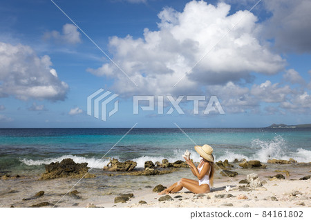 The girl in straw hat sitting on the beach with big seashell in the hands. 84161802