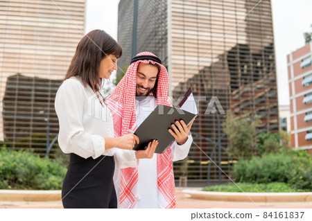 Smiling Arab man client singing contract after negotiation with european business woman while standing on city street, saudi businessman holding clipboard discussing deal details at outdoor meeting Smiling Arab man client singing contract after negotiation with european business woman while standing on city street, saudi businessman holding clipboard discussing deal details at outdoor meeting 84161837