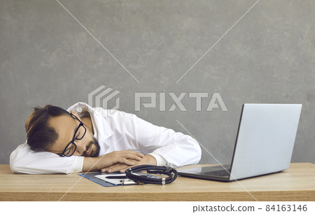 Tired young man in white coat sleeping sitting at office desk with stethoscope and laptop computer. Exhausted doctor or nurse dozes off after working on notebook PC. Lazy medical student lying on desk 84163146