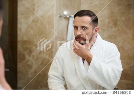 Handsome young European man in a white terry bathrobe touching the stubble before shaving, looking in surprise at his mirror reflection in the bathroom Handsome young European man in a white terry bathrobe touching the stubble before shaving, looking in surprise at his mirror reflection in the bathroom 84166808