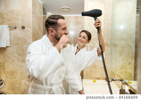 Happy middle aged loving couple having fun together at morning hygiene routine in bathroom. Handsome guy brushing teeth, smiles while his wife or girlfriend drying his hair using hair dryer Happy middle aged loving couple having fun together at morning hygiene routine in bathroom. Handsome guy brushing teeth, smiles while his wife or girlfriend drying his hair using hair dryer 84166809