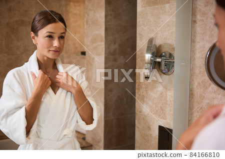 Beauty portrait of attractive middle aged European woman in white bathrobe looking at herself in the bathroom mirror, admiring herself and examining her face for wrinkles and the first signs of aging 84166810
