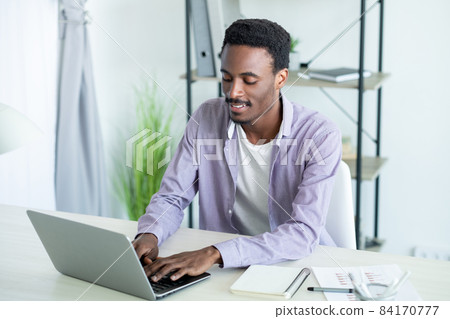 Home work. Busy man. Online communication. Concentrated smiling guy smart casual style typing laptop in light room interior. 84170777