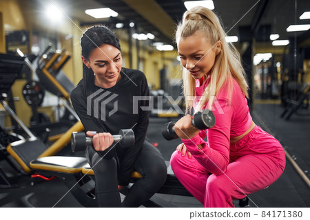 Female Coach shows the client an exercise for biceps with dumbbell while sitting on the bench in gym Female Coach shows the client an exercise for biceps with dumbbell while sitting on the bench in gym 84171380