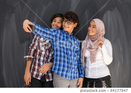 Group of arab teens taking selfie photo on smart phone with black chalkboard in background. Selective focus  84177847