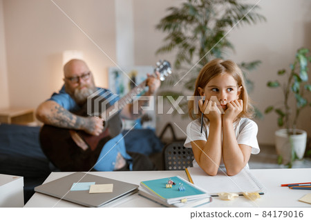 Schoolgirl sits at table while daddy bothers her playing guitar on sofa in living room 84179016