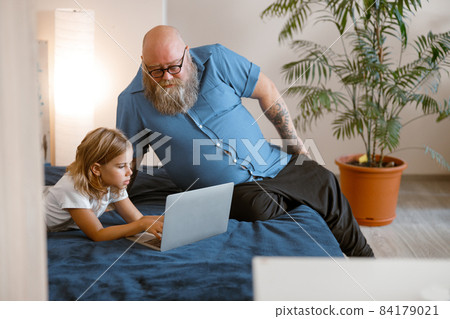Bearded plump man and attentive little girl watch video lesson via modern laptop on bed Bearded plump man and attentive little girl watch video lesson via modern laptop on bed 84179021