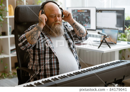 Bearded man listens to music with headphones sitting at sound synthesizer in home studio Bearded man listens to music with headphones sitting at sound synthesizer in home studio 84179041