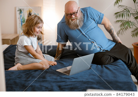 Bearded father and little girl watch moovie lesson via modern laptop sitting on bed Bearded father and little girl watch moovie lesson via modern laptop sitting on bed 84179043