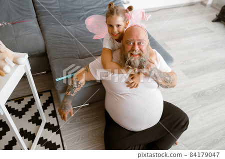 Happy little girl hugs funny obese daddy sitting on floor in living room Happy little girl hugs funny obese daddy sitting on floor in living room 84179047