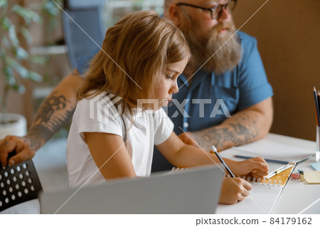 Pretty little girl writes in copybook sitting nera laptop doing homework with daddy at home 84179162
