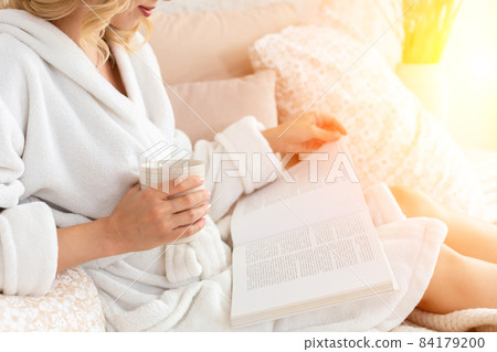 Young woman in white terry robe is drinking coffee and reading magazine or book in bedroom. Young woman in white terry robe is drinking coffee and reading magazine or book in bedroom. 84179200