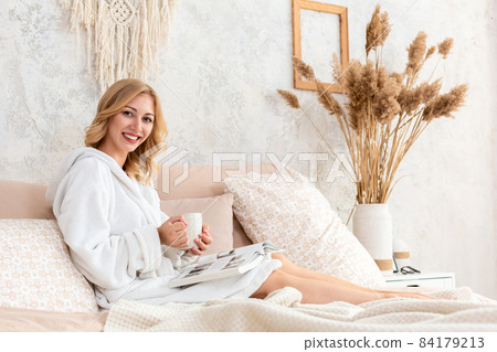 Young woman in white terry robe is drinking coffee and reading magazine or book in bedroom. Young woman in white terry robe is drinking coffee and reading magazine or book in bedroom. 84179213