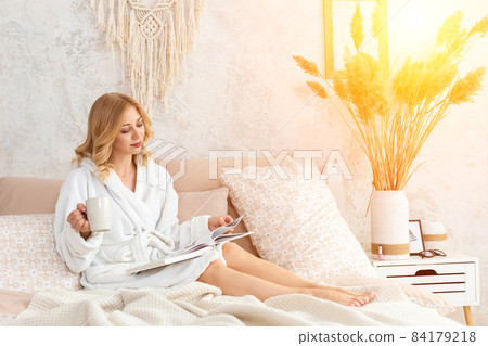 Young woman in white terry robe is drinking coffee and reading magazine or book in bedroom. Young woman in white terry robe is drinking coffee and reading magazine or book in bedroom. 84179218