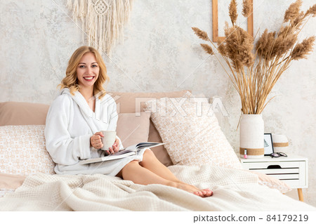 Young woman in white terry robe is drinking coffee and reading magazine or book in bedroom. Young woman in white terry robe is drinking coffee and reading magazine or book in bedroom. 84179219