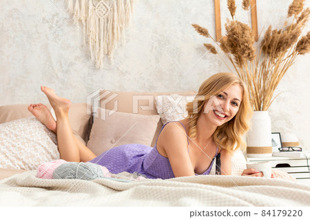Young woman holding a mug with tea or coffee sitting on the bed in the bedroom. Near lie yarn and knitting needles. Young woman holding a mug with tea or coffee sitting on the bed in the bedroom. Near lie yarn and knitting needles. 84179220