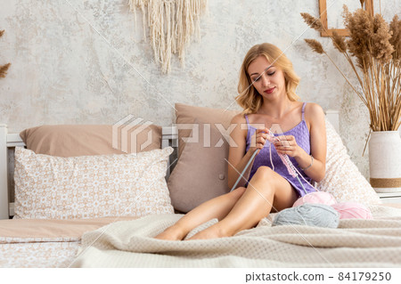 A young woman in a lilac home dress knits a light pink scarf or plaid from natural threads on the bed, against a white wall A young woman in a lilac home dress knits a light pink scarf or plaid from natural threads on the bed, against a white wall 84179250