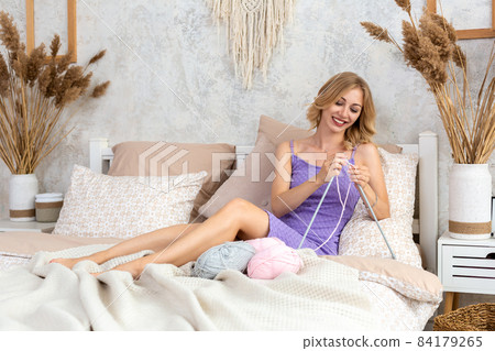 A young woman in a lilac home dress knits a light pink scarf or plaid from natural threads on the bed, against a white wall 84179265