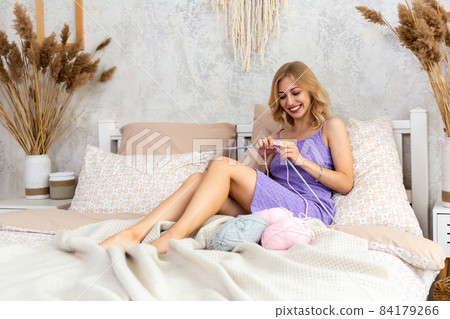 A young woman in a lilac home dress knits a light pink scarf or plaid from natural threads on the bed, against a white wall 84179266